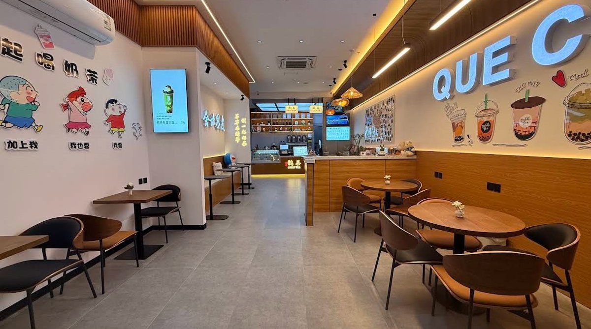 Interior of the Lucky Tea Ar Rabi Square cafe in Riyadh, with round wooden tables and chairs, a service counter, playful wall art, and warm pendant lighting